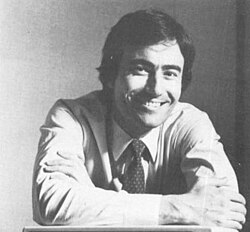 Grainy black and white portrait photo of a white man with dark hair smiling as he rests his arms on pedestal.