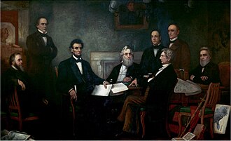 A dark-haired, bearded, middle-aged man holding documents is seated among seven other men.]]