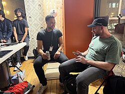 Two men sitting in folding chairs in a backstage area. One man wears a green shirt and a black baseball cap; the other wears a black t-shirt and holds a silver can. Wallpaper with various faces is visible in the background.