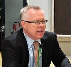 Photograph of Rob Monster from the shoulders up, wearing a suit and tie and speaking