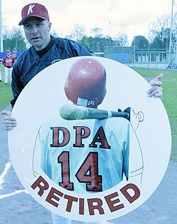Baseball player Marcel Joost holding a circular sign for his number retirement (showing the back of a baseball player with the number 14 and the word "retired" underneath)