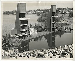 Black and white aerial photograph depicting a swimming pool surrounded by a seating area, and a stage with diving platforms on both ends
