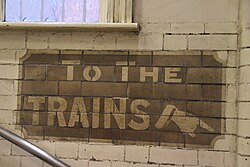A white-on-brown old sign on a staircase that reads "TO THE / TRAINS" with a manicule on the bottom right pointing down the staircase