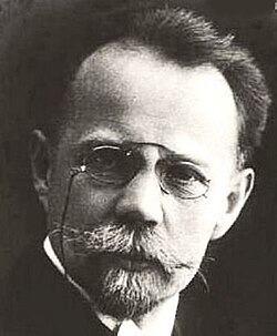 Black and white studio portrait of an unsmiling man wearing pince-nez, with short, dark hair brushed back, and a trimmed, greying beard.