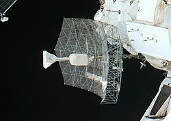 A rectangular dish shape of scaffolding covered in transparent sheeting, with a white insulation-covered radio receiver and support projecting from the centre. The blackness of space serves as the backdrop.