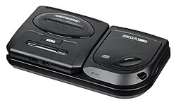 Photograph of an electronic video game console resting on a compact disc attachment. The console is encased in black matte plastic that is shaped as a flattened rectangular cuboid with rounded corners. The top has two buttons and a slot for cartridges, while the front has two ports. The attachment as a top-loading disc tray with a button labeled "open".