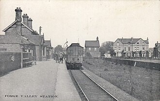 A black and white image of a train static in a station, with the platform on the left and houses beyond on the right