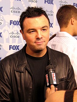 A man with dark hair, sporting a leather jacket, and being interviewed. There is a small microphone in front of him, with a television channel logo placed onto it.