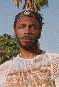 Hendricks stands in front of palm trees and stage equipment in the bright sun, wearing sturdy boots.