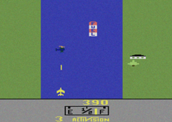 A rectangular video game screenshot that is a digital representation of a jet fighter over a river and two grass-filled shores. The jet fighter is shown firing a missile at a helicopter while a tree and a dwelling are displayed on the grassy portion. A tank labeled "Fuel" is shown resting on the water.