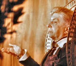 A sepia-toned photograph. A straight-faced Quentin Crisp gestures from an ornate, high-backed chair. A large, red handkerchief flops from her jacket pocket.