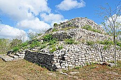 A tourist’s photo: angled towards us is a pyramid-like Mayan structure made out of stones with grass growing out of the stones. The sky is blue with clouds on the left.