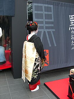 A maiko with the sakko hairstyle who is dressed in a black hikizuri and gold brocade obi tied in the darari knot, is posed with her back towards the camera in order to show her hairstyle and obi design.