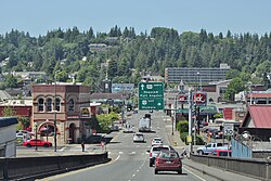 Looking down from a one-way bridge with two lanes at a city skyline with low-slung buildings and hills in the background; a sign reading "101 North Hoquiam, Port Angeles; 12 East Olympia" hangs over the highway.