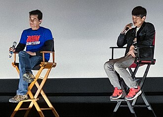 Photograph of two men seated in film director's chairs in front of a projection screen