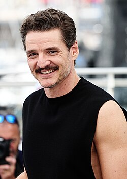 A smiling man with brown hair, brown facial hair, wearing a green jacket and blue-and-white striped shirt, smiling at something off-camera.
