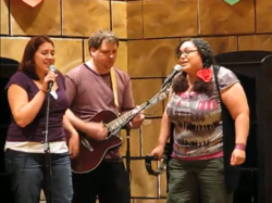 Three people are standing in front of a papered wall that has been made to appear as the interior of a castle with tan stonework. There are two fireplaces visible, one on either side of the corner behind them. Banners are hanging on the wall, with only the very bottoms visible in frame. The person on the left is singing into a microphone; the person in the middle, standing slightly behind the other two, is playing guitar; the person on the right is also singing into a microphone but is also playing solo tambourine, without the skin stretched over one side of the instrument.
