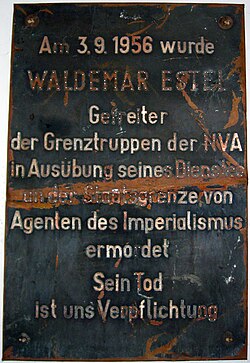 Damaged metal plaque reading "Am 3.9.1956 wurde WALDEMAR ESTEL Getreiter der Grenztruppen der NVA in Ausübung seine Dienstes an der Staatsgrenze von Agenten des Imperialismus ermordet. Sein Tod ist uns Verpflichtung."