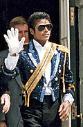 Michael Jackson, with sunglasses, curly hair, and diamond-encrusted clothing, waves with his rhinestone-covered white glove as a man stands behind him.