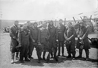 Nine men wearing a mixture of military uniforms with caps and flying suits with goggles, in front of a row of military biplanes