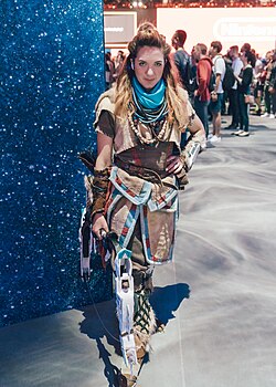 A woman in detailed cosplay as Aloy. She has red braided hair and is wearing a textured outfit made of leather, fur, and beads with a prominent blue scarf