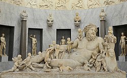 A large marble sculpture, inside a formal room. The main object is a reclining nude man, surrounded by many children, as well as some animals.