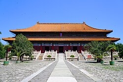 Front view of a large traditional Chinese palace building with a yellow-tiled roof, centered along a stone pathway and stairway, surrounded by trees under a clear blue sky