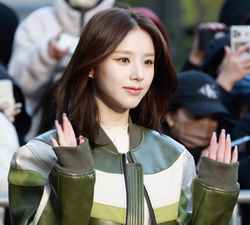 A young Asian woman with shoulder length brown hair and brown eyes wearing a green leather jacket waving with both of her hands.