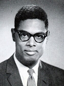 A dark-haired man, wearing glasses and a suit and tie, looks into the camera