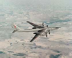 Top perpendicular starboard view of four-engine propeller-driven aircraft in-flight. The pale gray/white aircraft is flying above almost white terrain.