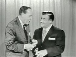 two men stand in front of a curtain speaking to each other