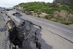 The remains of an asphalt road that has been destroyed with several pieces lifted up from the ground and others with large cracks.