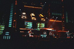 An elaborate concert stage set, set in a dark stadium. Three automobiles hang above two projector screens, aiming their headlights towards the stage.