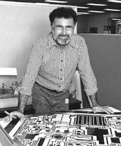 A man with a beard leans over a table containing technical drawings of an electronic circuit