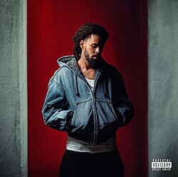 J. Cole standing in front of a wall while looking downwards with a somber expression and resting his hands in his jacket's front pockets