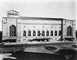 Black and white external image of a multiple-storey brick building with large archways for doors, and smaller archways for windows