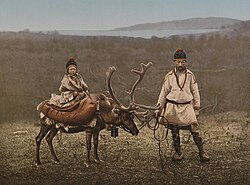 A historic photograph of a Sámi man standing beside a reindeer with a child seated on the animal, taken in Finnmark, northern Norway, around 1890–1900.