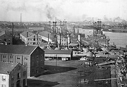 Several World War I ships line a port crowded with warehouses, with a city skyline behind them.