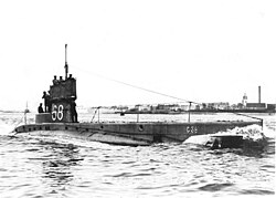 Black and white photograph of a C-class submarine surfacing in port. Five men are visible standing on the rear of the vessel