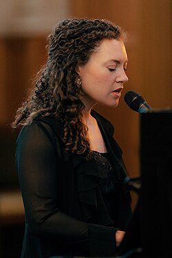 close-up of Celine Cairo wearing a black top, playing piano and appearing to sing into a microphone with eyes partially closed