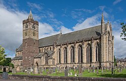 Side view of the nave of a cathedral from outside. Tall arched glass windows run along half the length of the nave from the right. Adjacent to the nave, and to the left of the scene is a cuboid-shaped tower with a conical spire. The foreground is scattered with headstones of a graveyard on green grass.