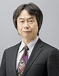 alt=Portrait photo of a Japanese man with black hair wearing a black business suit and a colorful tie.