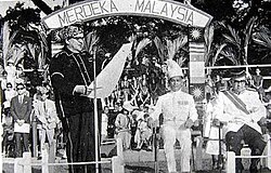 A man reading from a large piece of paper in front of an arch saying "MERDEKA MALAYSIA"