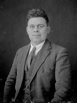 "Black and white photograph of a man aged c. 30 with dark hair swept upwards above his forehead; he is wearing a white shirt and tie and a three-piece suit"
