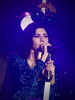 A woman singing, dressed in sparkling blue dress with a glittery cherry headpiece, under purple lighting.