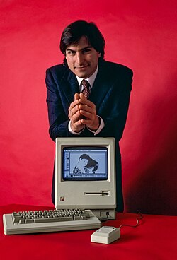 With a red background, Steve Jobs rests his forearms on a Macintosh computer.