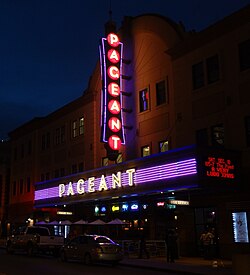 Photographed from an oblique angle, the Pageant's façade is lit by two perpendicular signs (in purple, yellow, and red) displaying its name, as well as a small marquee advertisement