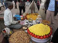 A street food vendor spoons onions onto newsprint to serve. His stall has several ingredients, including puffed rice, peanuts, tomatoes, lemons, and chillis.