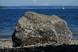 A boulder sits on a cobbled beach in bright sunlight with a stretch of blue water behind it. On the horizon are low forested hills. There is a snow covered volcano on the far distance. A few sailboats are in the water.