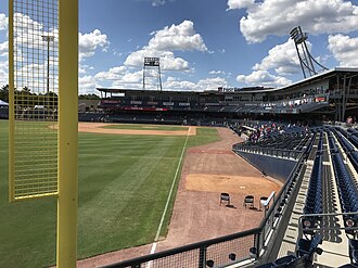 A green baseball field surrounded by its brown shale warning track and blue seats viewed from the concourse by the yellow left field foul pole on a sunny afternoon